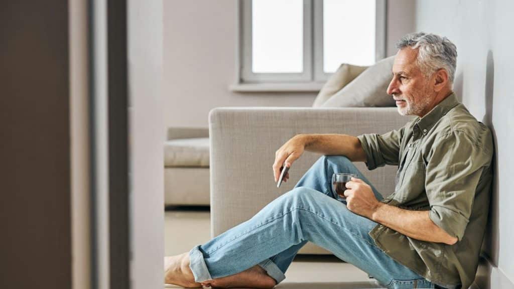 Man with gray hair and beard sits on floor, holding a cup and smartphone.