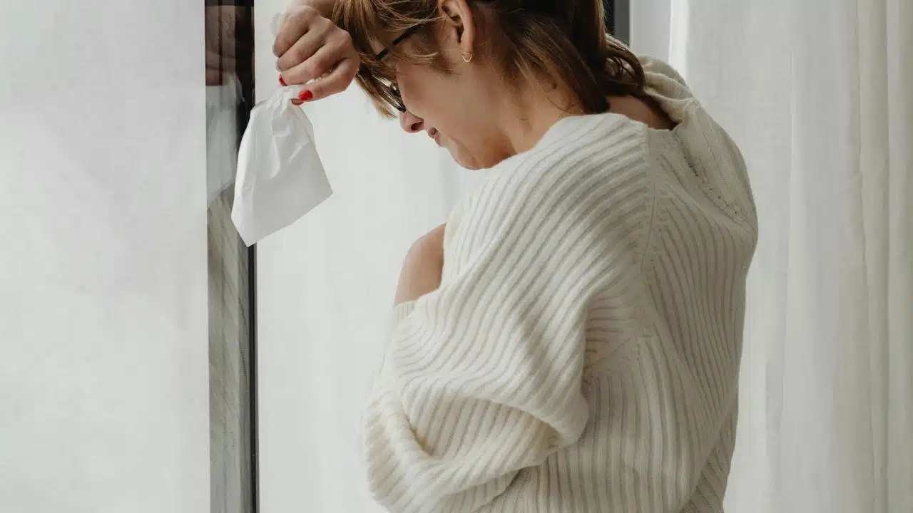 A woman holding a tissue and leaning against a window.