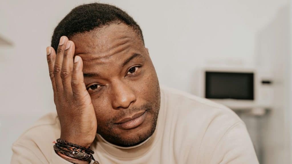 African-American man in a tan shirt holds his hand to his head, looking troubled.