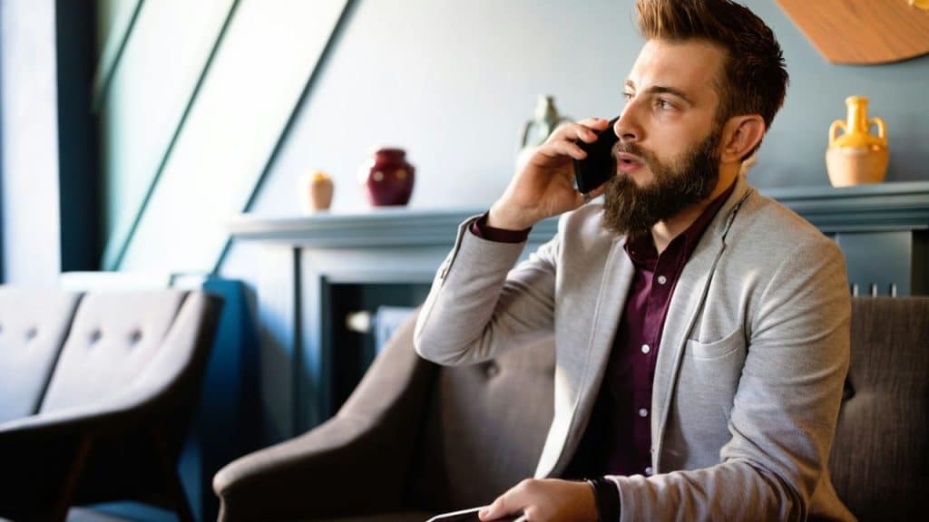 Man with a beard and gray jacket talking on a cell phone in a cafe.