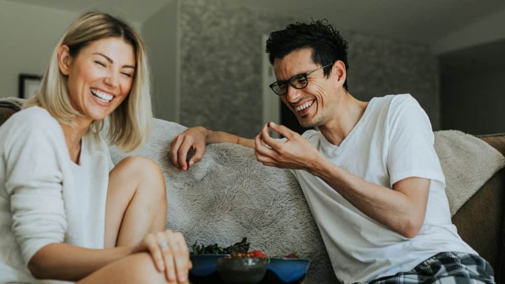A man and woman laugh while sitting on a couch and eating snacks from bowls.