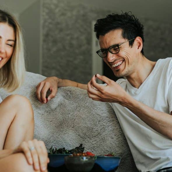 A man and woman laugh while sitting on a couch and eating snacks from bowls.
