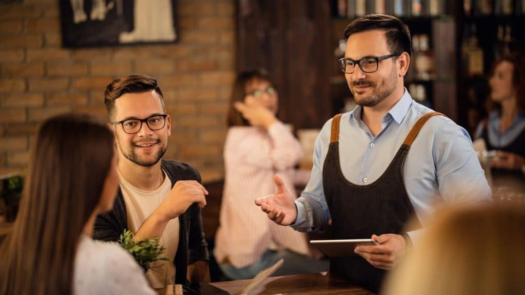 A man in glasses and an apron stands at a table holding a digital tablet.