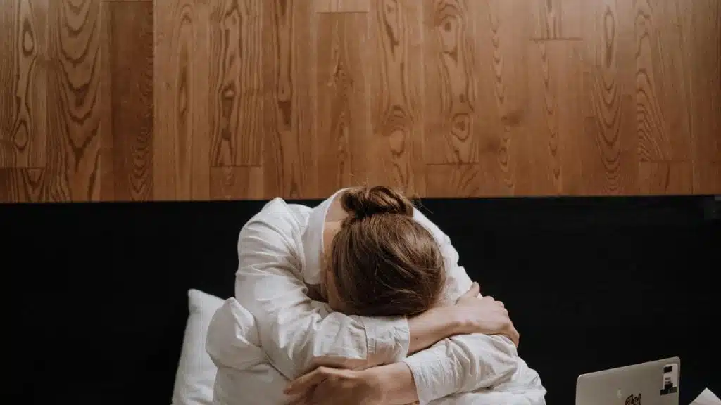 A person sitting on a bed with their head down and arms wrapped around their knees.