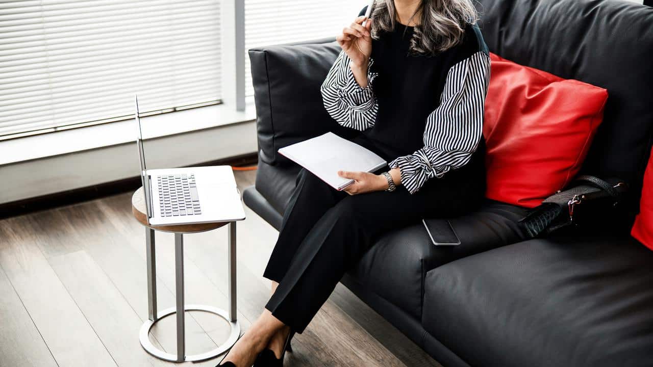 A woman sitting on a couch with a laptop and notebook, appearing focused.