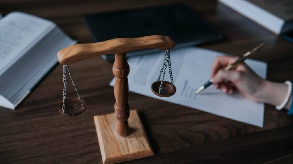 A wooden scale of justice on a desk with a person writing on legal documents.