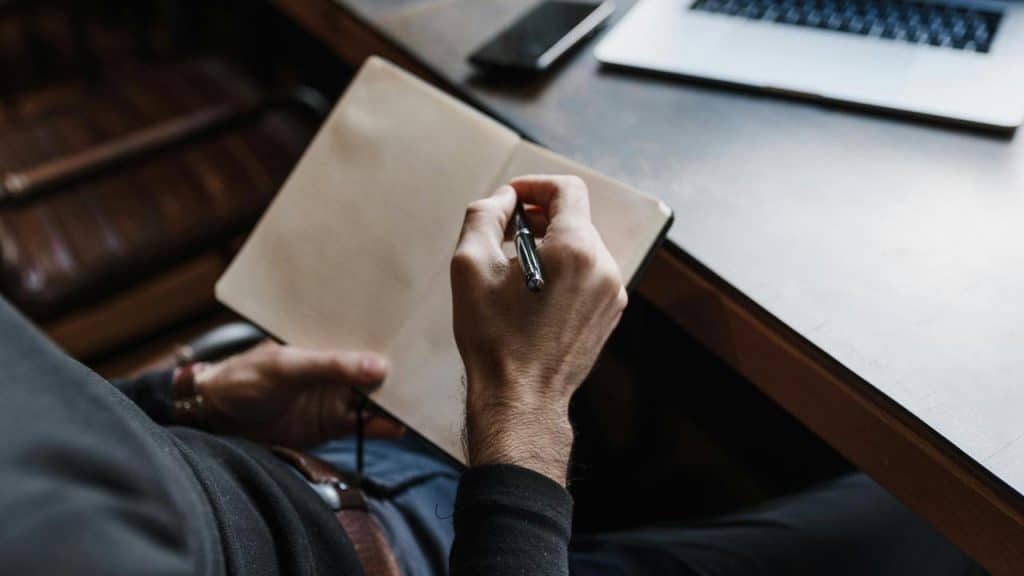 A person writing in a blank notebook at a desk with a laptop nearby.