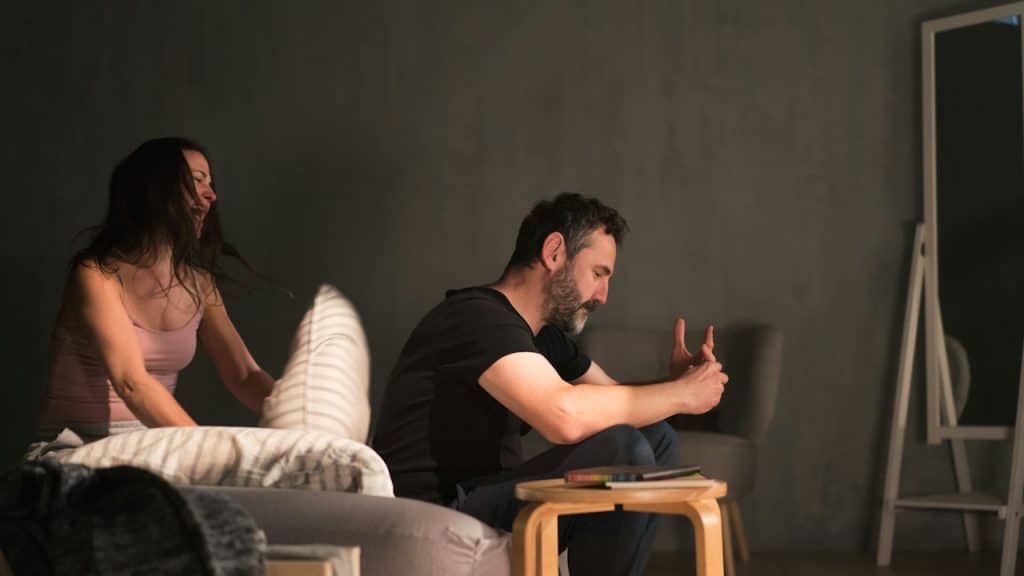 A man is sitting with his hands clasped, looking stressed or upset, while a woman beside him appears to be throwing a pillow.