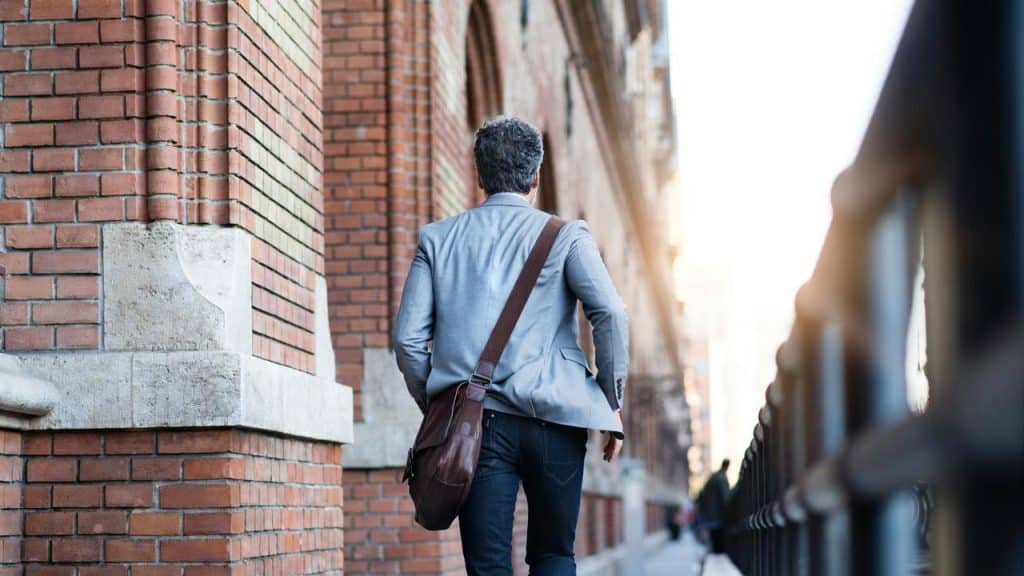 A man walking along a brick building with a shoulder bag.