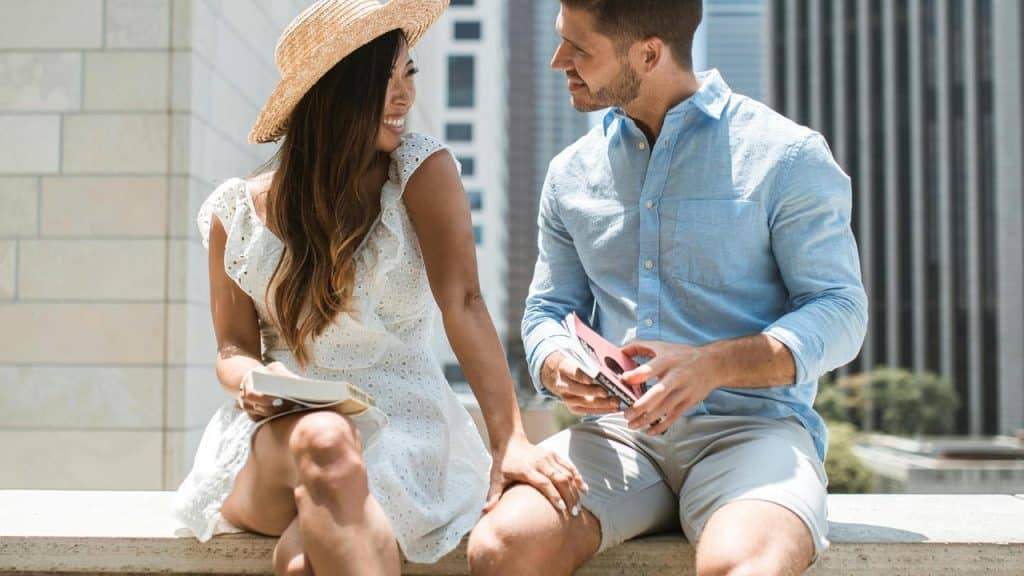 A couple sitting outdoors on a ledge smiling at each other.