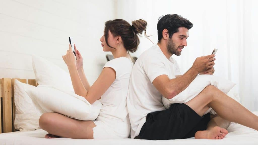 A man and woman sit back-to-back on a bed while looking at their smartphones.