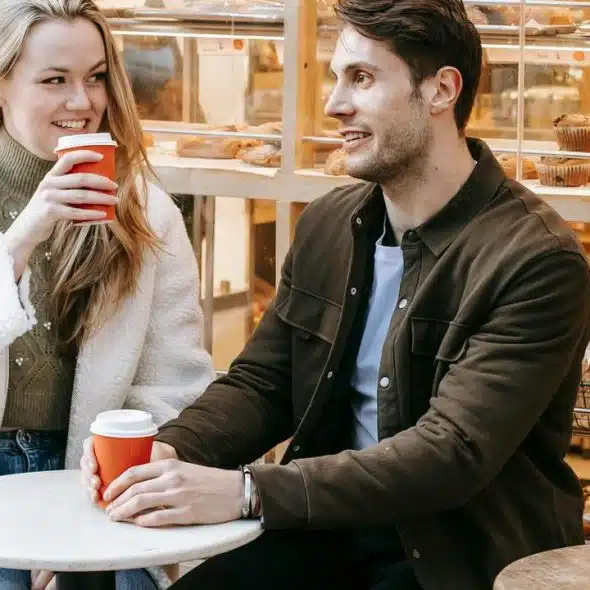 A man and woman sitting at an outdoor café table, smiling and holding coffee cups.