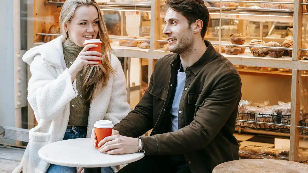 A man and woman sitting at an outdoor café table, smiling and holding coffee cups.