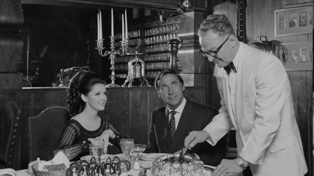 A waiter cutting a cake for a couple sitting at a dinner table.