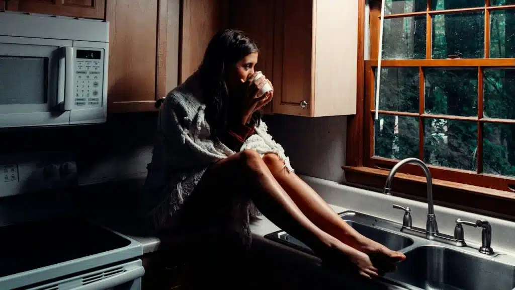 A woman drinking coffee while sitting on a kitchen sink.