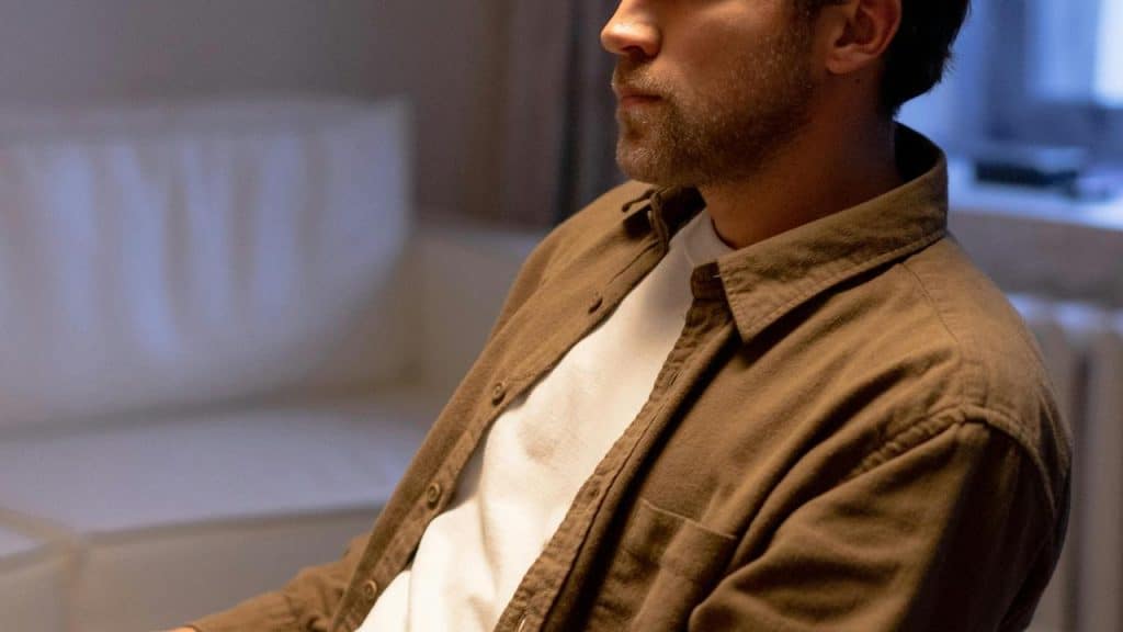 A man in a brown shirt sits indoors in soft lighting.