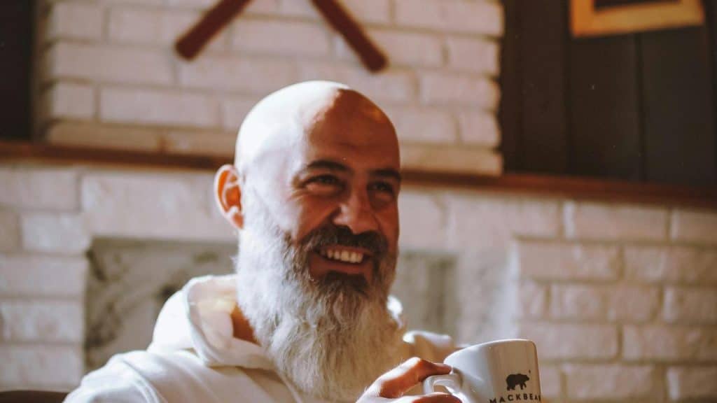 A bearded man smiling while holding a coffee mug indoors.
