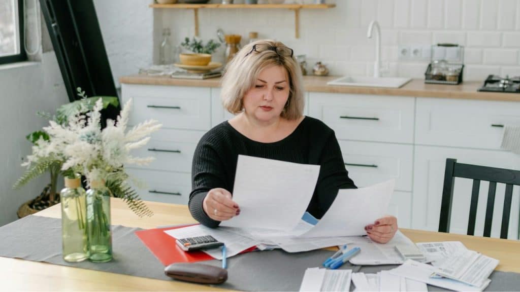 Woman with blonde hair reading papers at a kitchen table with a calculator.