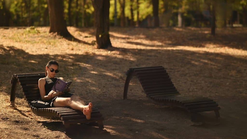 A woman wearing sunglasses and a dark outfit is lying on a wooden lounge chair, reading a book in a forest area.
