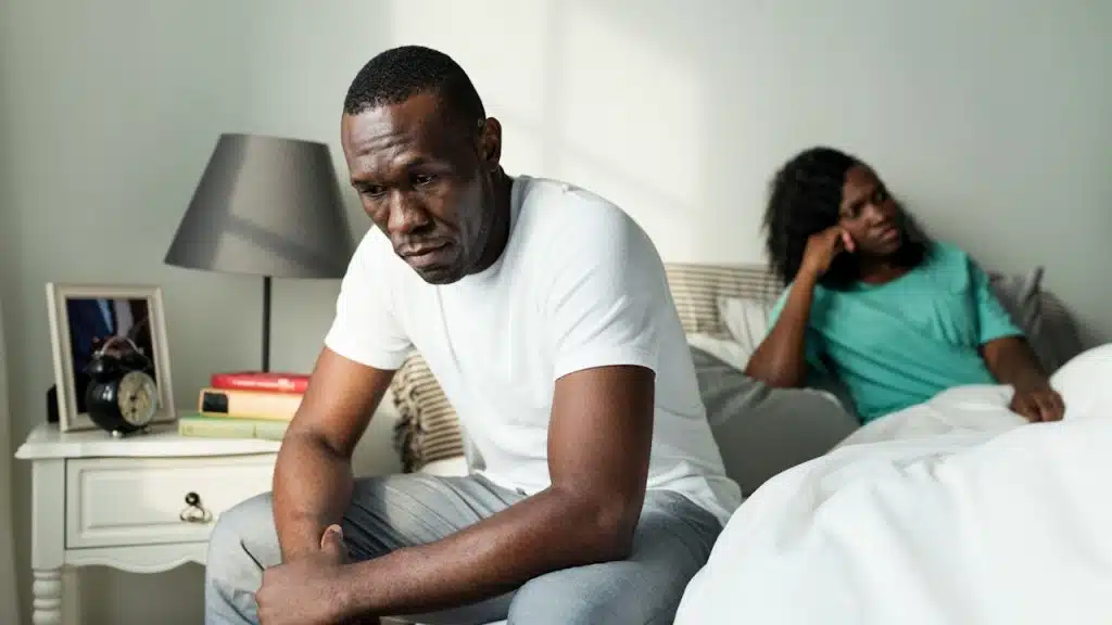 Man sitting on the edge of a bed looking distressed, with a woman sitting in the background facing away from him.