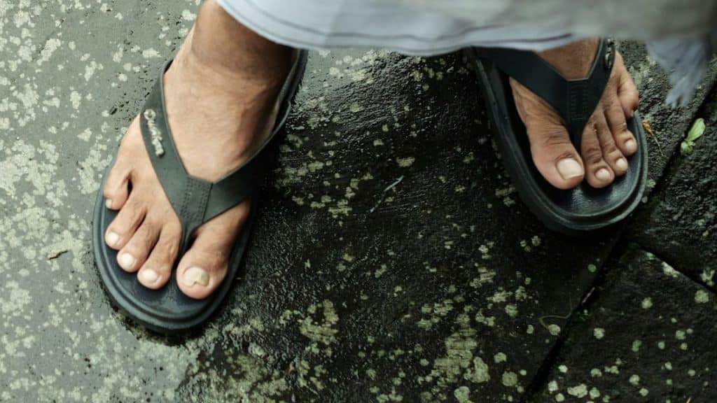 Overhead view of bare feet wearing black flip-flops on wet, dark, textured pavement.