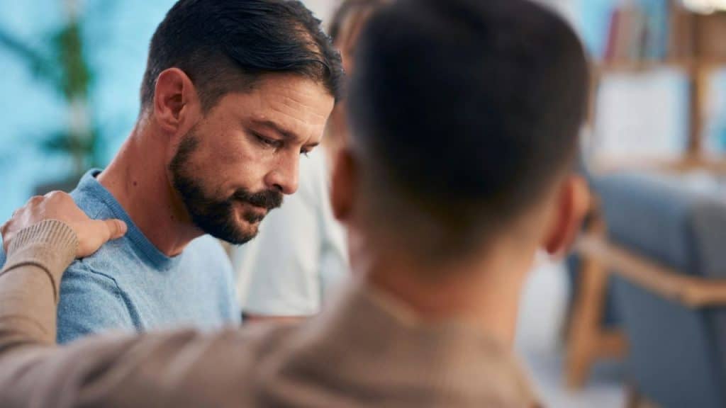 Bearded man in a blue shirt looks down while another man places a comforting hand on his shoulder.