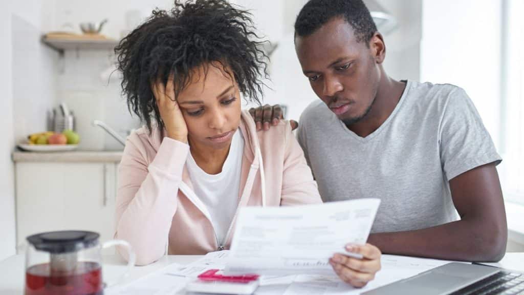 Two people looking stressed while reviewing papers and a calculator at a kitchen table.