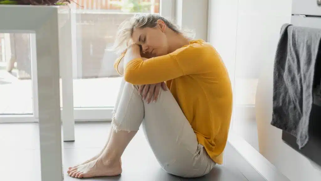 Woman in a yellow top and white pants sitting alone on the floor by a window.