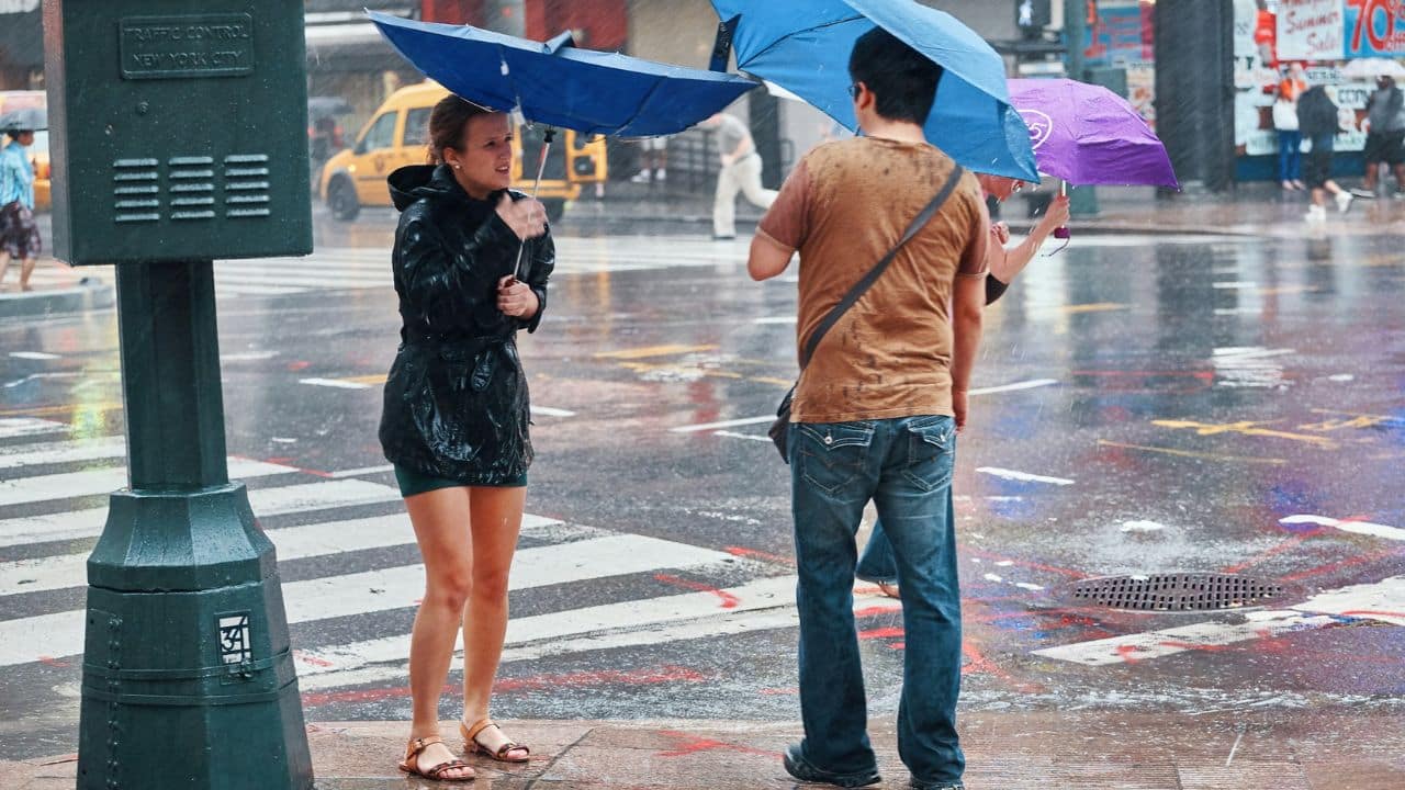 Three people stand in the heavy rain in a city street, two holding blue umbrellas.