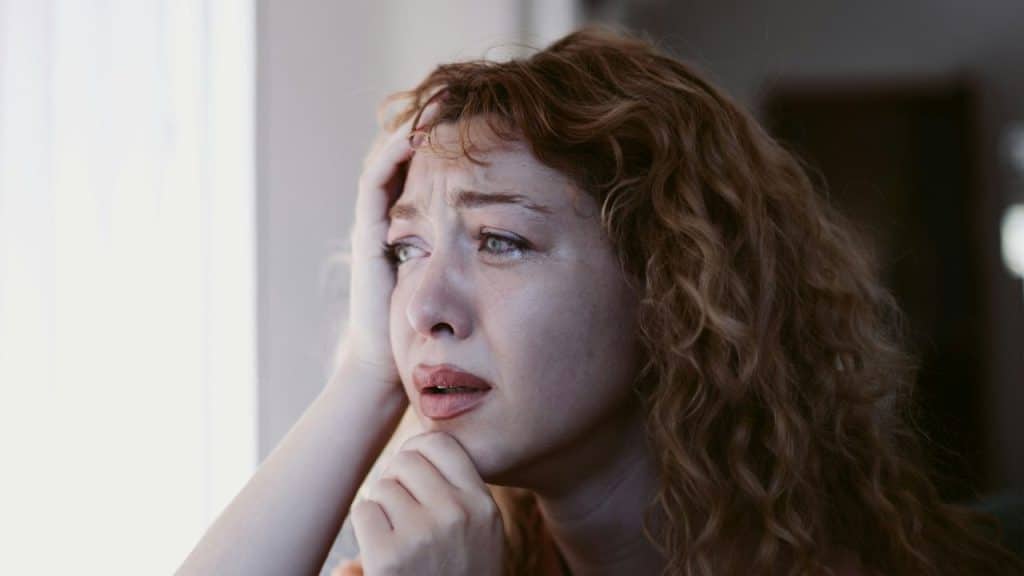 A close-up of a distraught woman with red hair and tears on her face, holding her head.