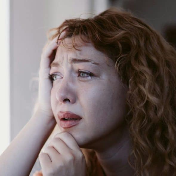 A close-up of a distraught woman with red hair and tears on her face, holding her head.