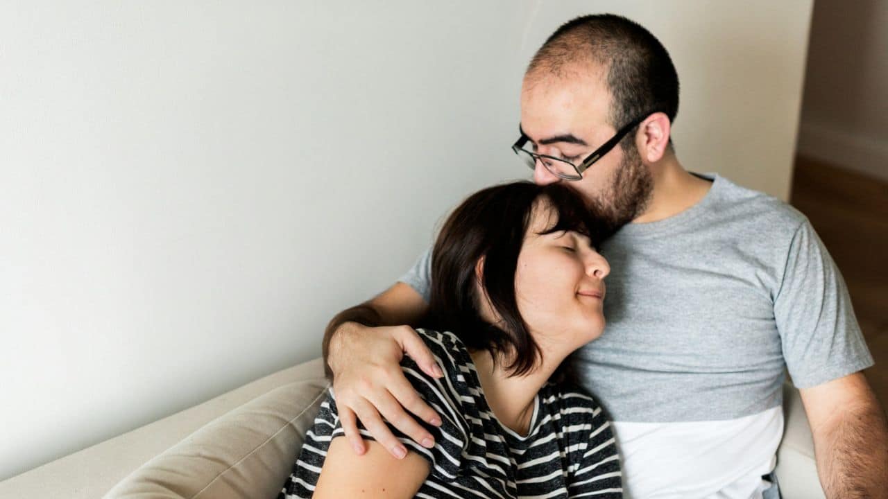 Man wearing glasses kissing the forehead of a woman leaning into his chest.