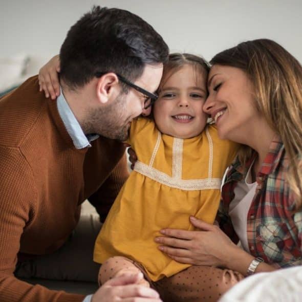 Happy family sitting on the floor, embracing their young daughter who is smiling at the camera.