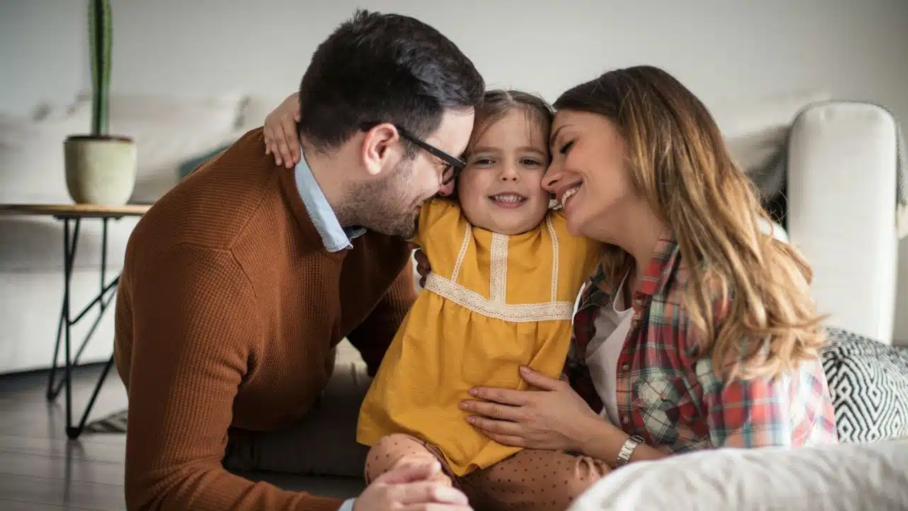 Happy family sitting on the floor, embracing their young daughter who is smiling at the camera.