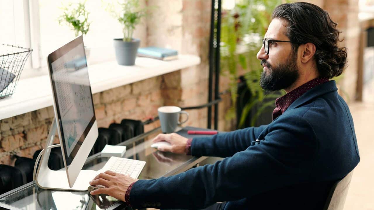 Man with a beard and glasses working at a computer in a home office setting.