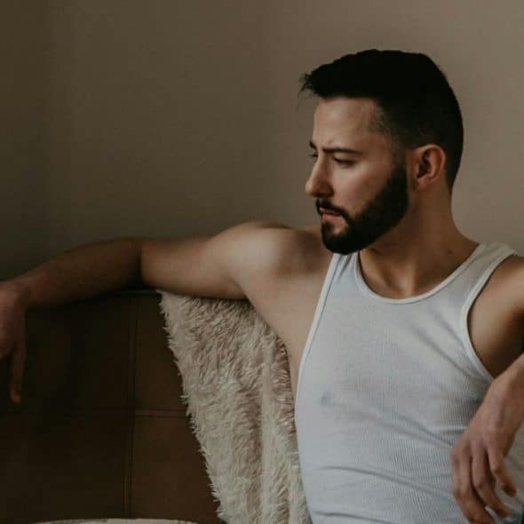 A bearded man in a white tank top sits looking downcast against a wall.