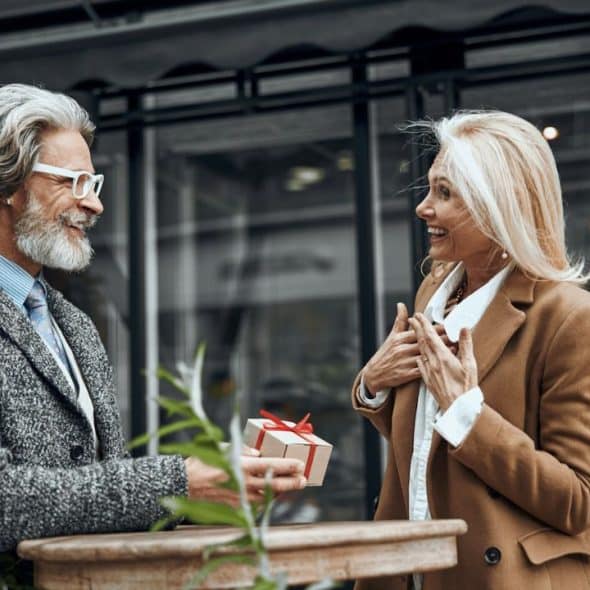 Mature man presenting a small gift box to a surprised woman outdoors near a cafe.