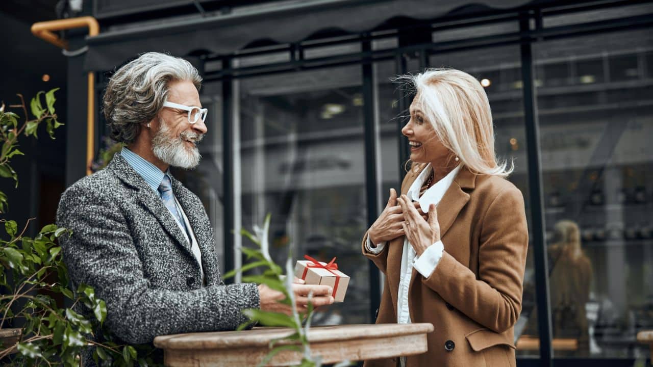 Mature man presenting a small gift box to a surprised woman outdoors near a cafe.