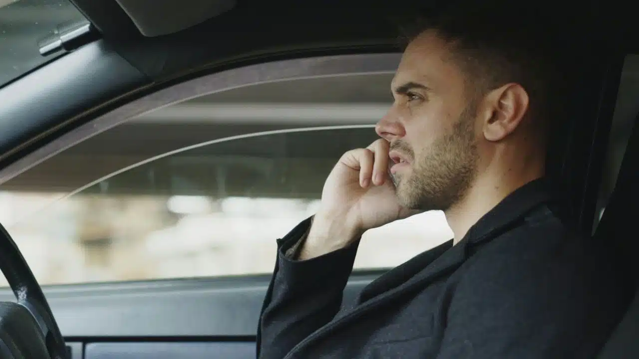 A man in a black coat sits in a car, holding a phone to his ear, looking serious or concerned.