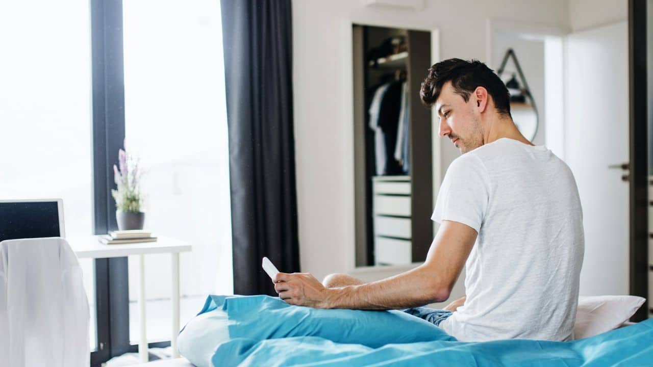 A man in a white t-shirt sits on a bed, looking down at his phone.