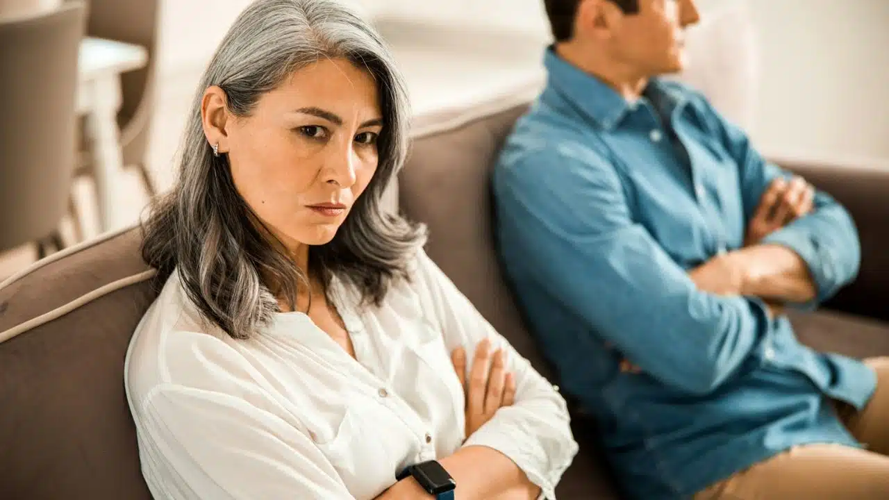 A woman with arms crossed sits on a couch, looking angry, with a man behind her.
