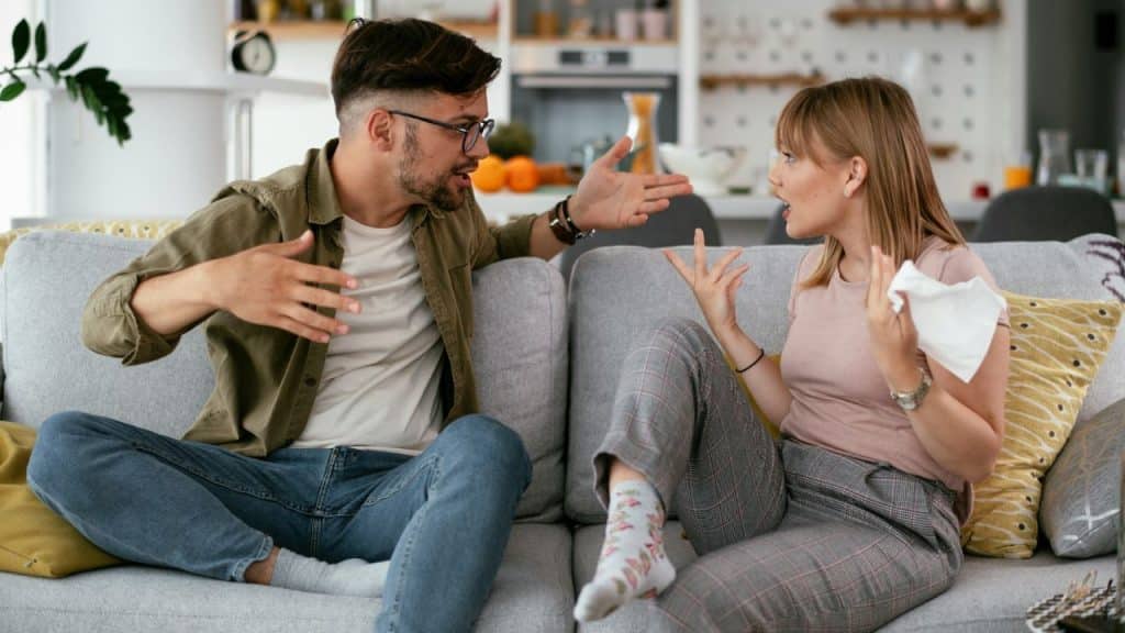 A man and woman sit on a sofa, gesturing expressively during a heated discussion.