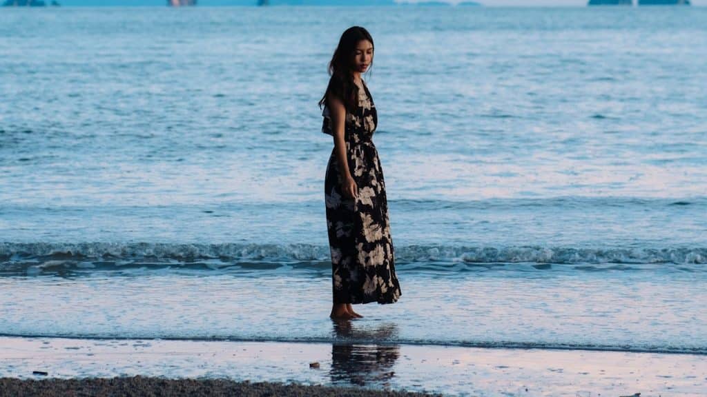 A young woman wearing a long black dress is standing on a beach next to the ocean.