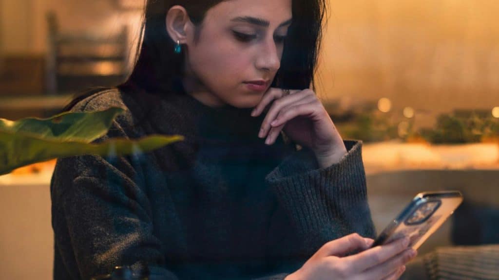 A woman sits indoors, thoughtfully looking at her phone in warm light.