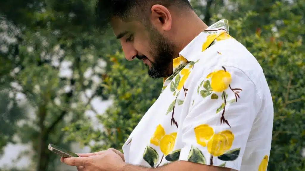 A bearded man wearing a patterned shirt and texting on his phone while standing outdoors.