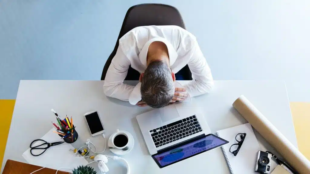 A man slumped over his desk with his face in his arms, surrounded by work supplies.