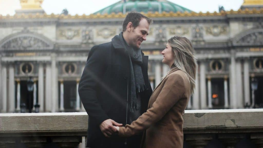 Couple in winter coats holding hands in front of a grand European building.
