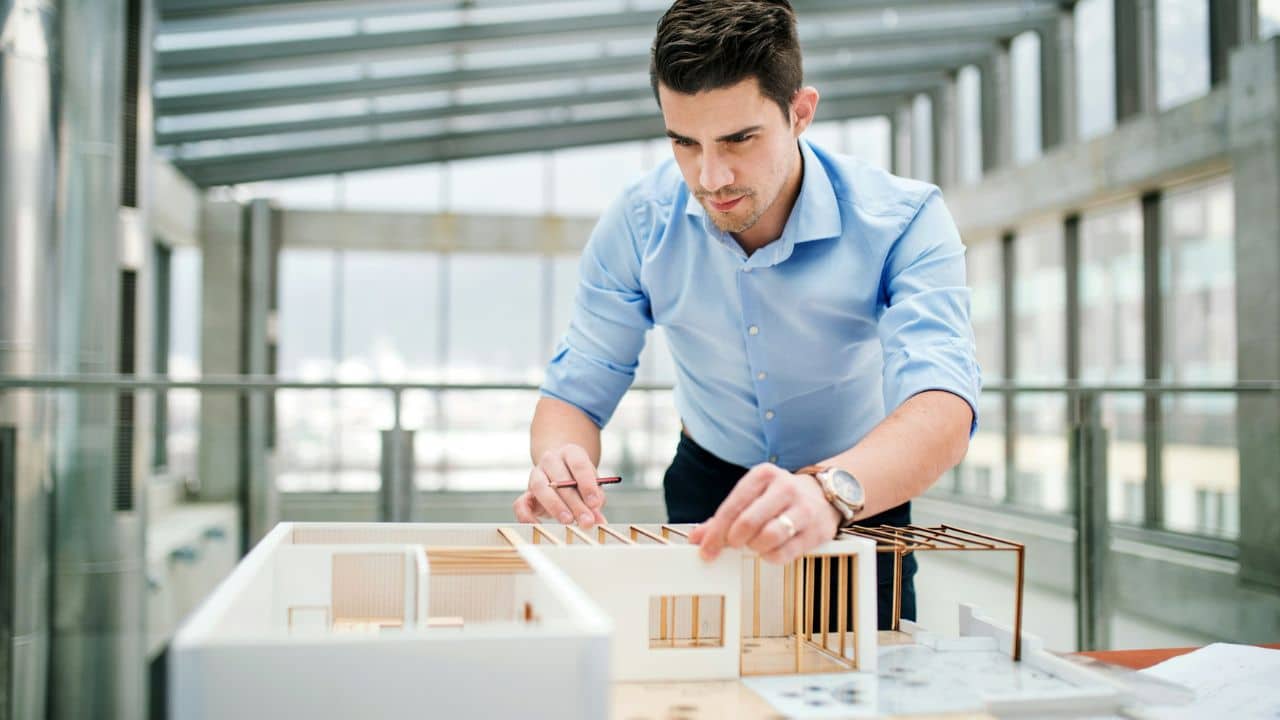 A man in a blue shirt and dark pants is working on a white scale model of a house in a modern interior space.