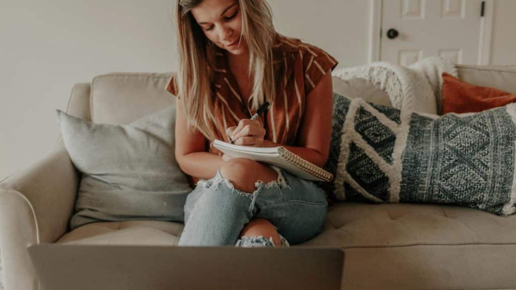 A woman sitting on a couch taking notes with a laptop in front of her.