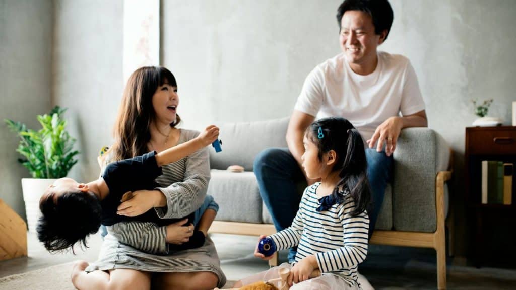 A family of four sits on the floor and a couch, smiling and playing together indoors.